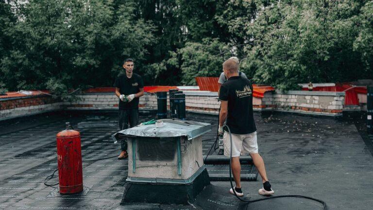 Two workers are performing maintenance on a flat roof with equipment and tools.